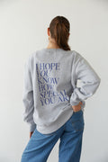 A person wearing a gray crewneck sweatshirt with text on the back standing against a white background.