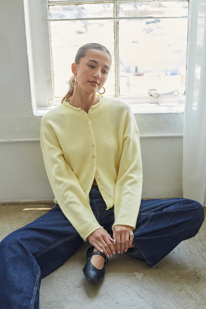 Person wearing a light yellow cardigan and blue jeans sitting on the floor in front of a window.