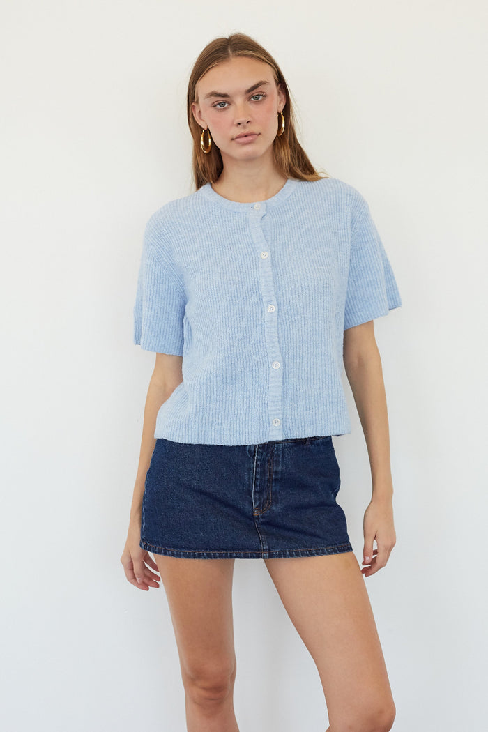 A woman modeling a light blue short-sleeve button-front knit top with a dark blue denim mini skirt, standing against a plain white background.