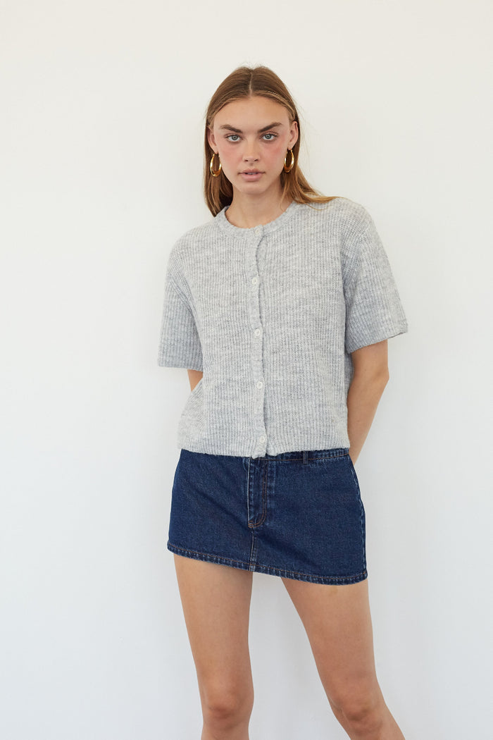 A woman modeling a light gray short-sleeve textured button-front top with a dark blue denim mini skirt, standing against a plain white wall.
