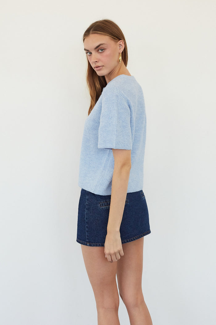 A woman modeling a light blue short-sleeve knit top and dark denim shorts against a plain white studio background.