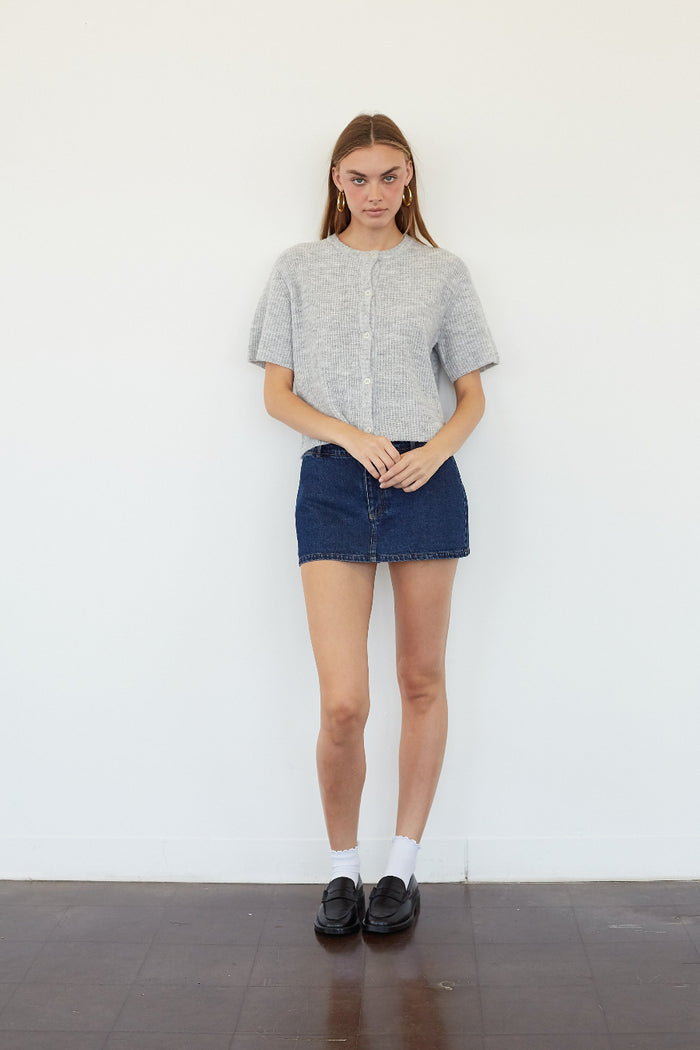A woman stands against a white wall wearing a light gray short-sleeve knit top, a dark blue denim mini skirt, white socks, and black loafers.