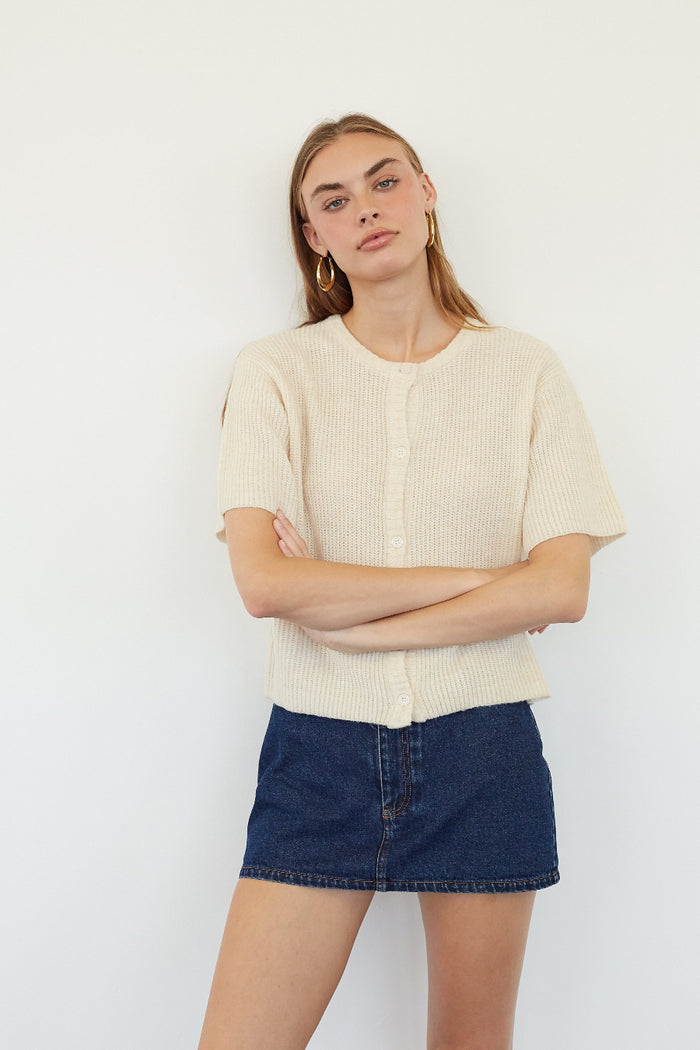 A woman modeling a cream ribbed short-sleeve cropped cardigan with a button placket, paired with a dark blue denim mini skirt, against a plain white studio background.