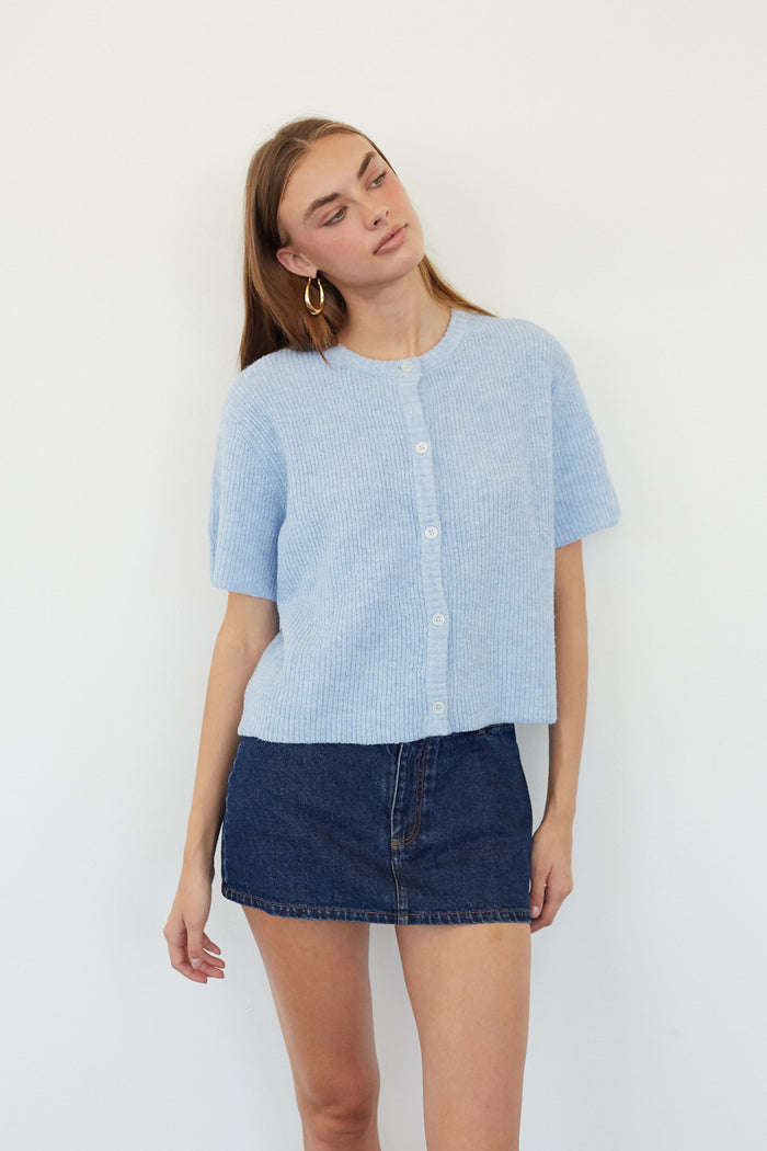 A woman modeling a light blue short-sleeve textured knit top with white buttons, paired with a dark denim mini skirt, against a plain white background.