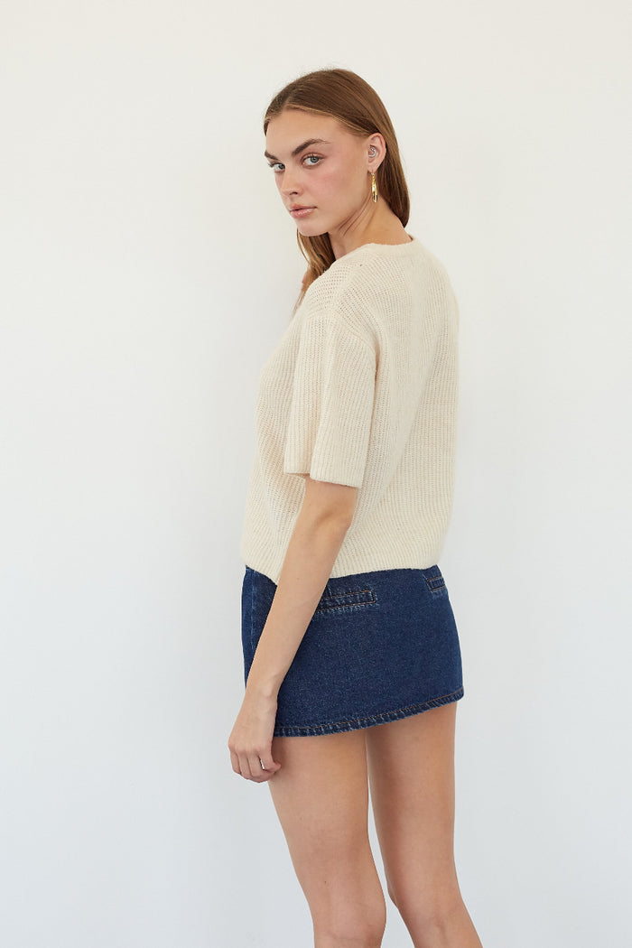 A woman modeling a cream-colored short-sleeve ribbed knit top and a dark blue denim mini skirt against a plain white studio background.