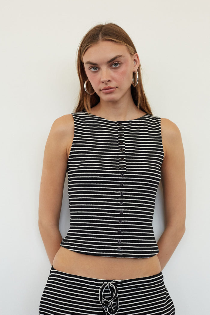 A woman modeling a sleeveless black and white horizontally striped crop top with a button-down front, paired with matching striped bottoms, against a plain white background.