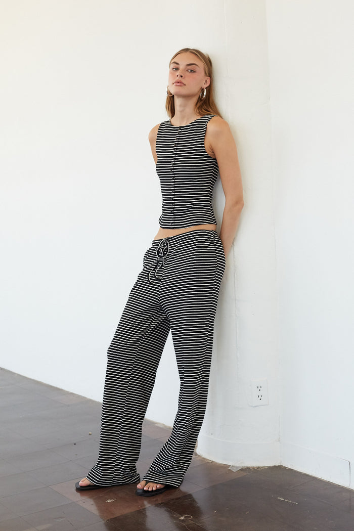 A woman stands against a white wall in a minimalist room with dark wood floors, wearing a matching black and white striped sleeveless crop top and wide-leg pants.