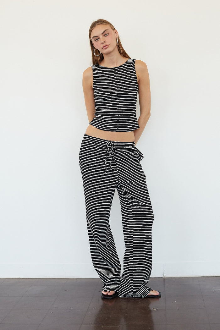 A woman modeling a matching black and white striped two-piece set consisting of a sleeveless cropped top with buttons and wide-leg drawstring pants, standing against a white wall.