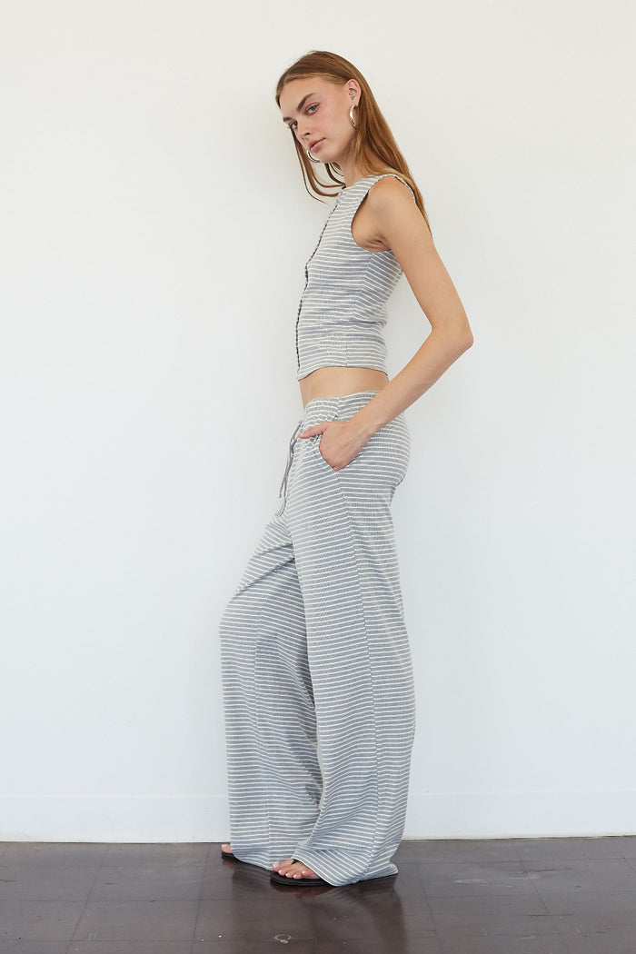 A woman modeling a matching two-piece loungewear set consisting of a sleeveless crop top and wide-leg pants, both in a light gray and white striped pattern, standing against a plain white wall.