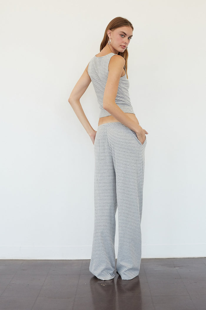 A woman modeling a matching two-piece loungewear set consisting of a sleeveless top and wide-leg pants, both in a light gray and white striped pattern, standing against a plain white wall.
