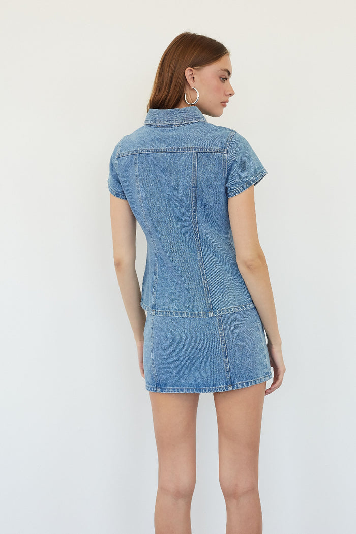 A woman shown from the back wearing a light blue denim short-sleeve mini dress with a button-down collar, photographed against a plain white studio background.
