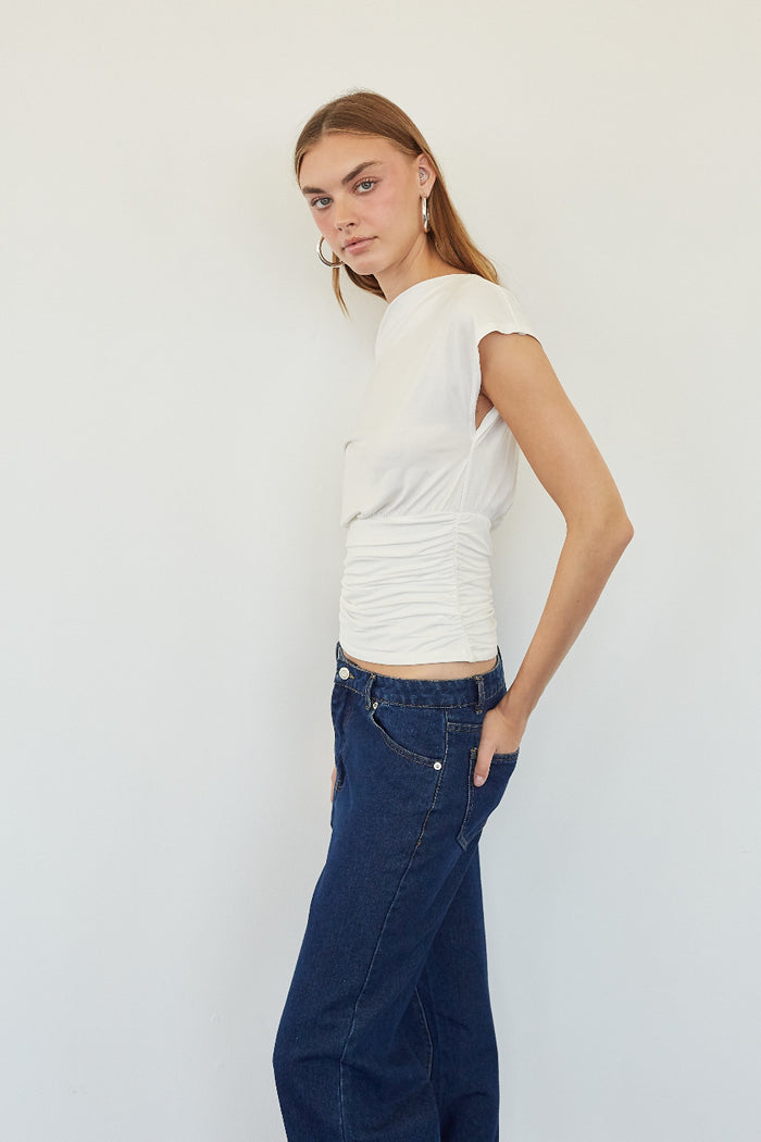 A woman modeling a white short-sleeve top with ruched detailing at the waist, paired with dark blue jeans, against a plain light gray background.