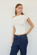 A woman modeling a white off-the-shoulder short-sleeve top with ruched detailing, paired with dark blue jeans, against a plain white wall.