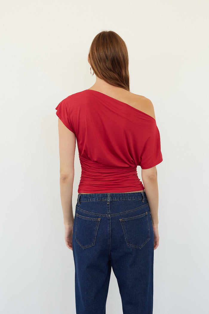 A woman viewed from the back wearing a bright red one-shoulder top and dark blue jeans against a plain white background.