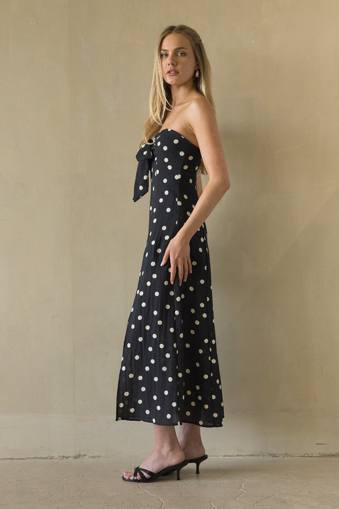 Woman wearing a black strapless dress with white polka dots against a beige wall.
