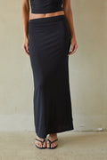 A woman wearing a long black wrap-style maxi skirt with a matching black crop top, standing against a textured beige wall.