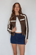 Woman wearing a brown jacket with white stripes, white top, and blue skirt on a plain background
