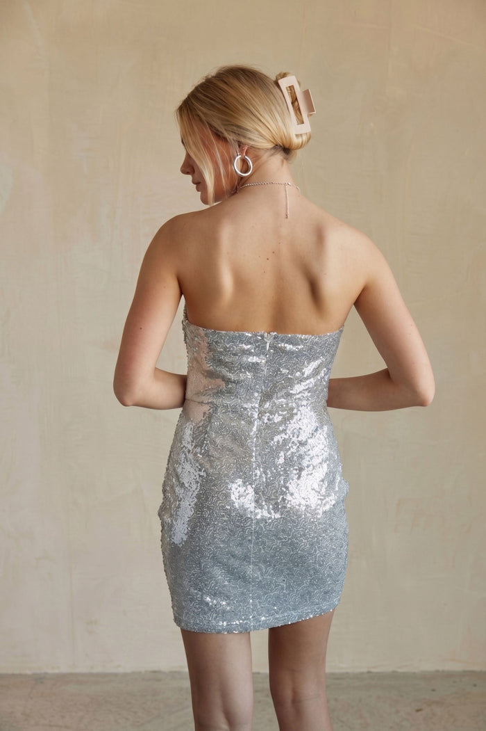 A woman shown from the back wearing a strapless silver sequin mini dress against a neutral beige textured wall.