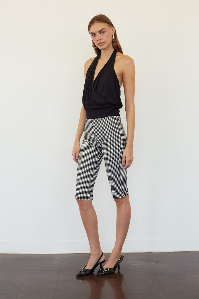 A woman modeling black and white gingham checkered capri pants with a black halter top, standing against a plain white wall.