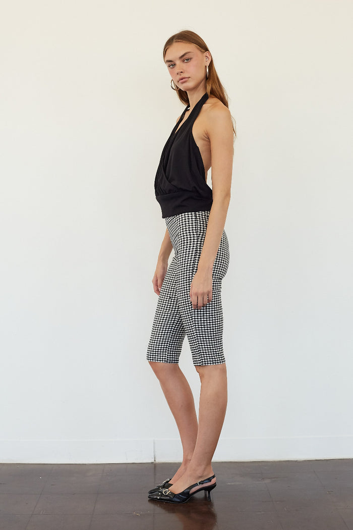 A woman stands in profile wearing a black halter top and black-and-white gingham biker shorts against a plain white wall.