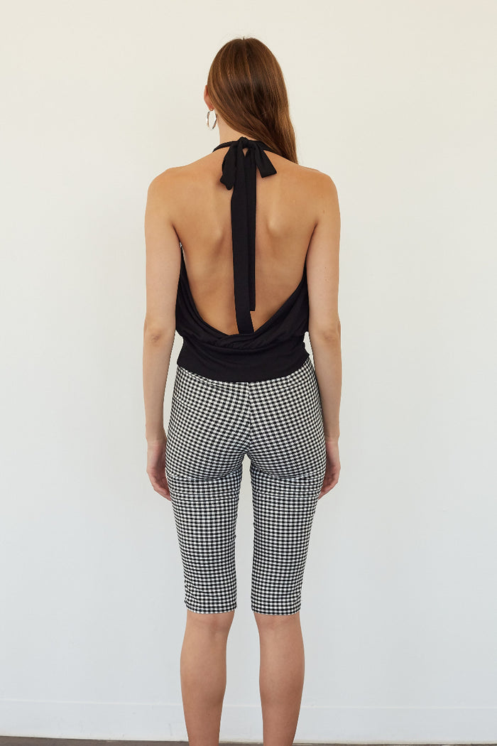 A woman shown from the back wearing a black halter-style top with a tie-back and black-and-white gingham capri pants, standing against a plain white wall.