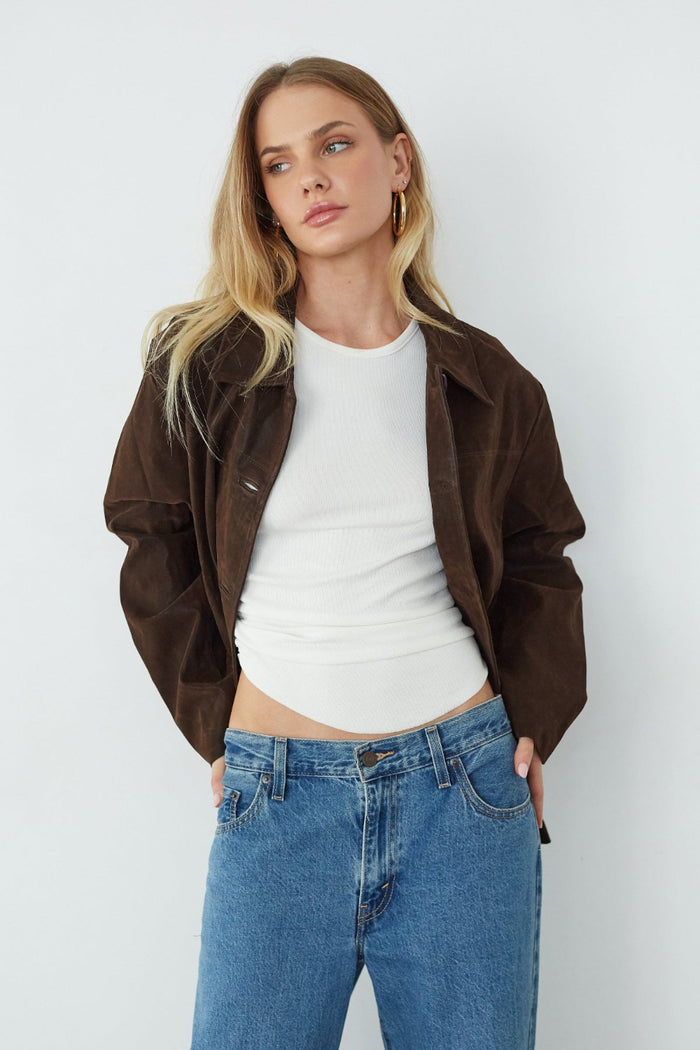 Woman wearing a brown jacket, white shirt, and blue jeans against a white background
