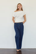 A woman stands against a plain white wall wearing a white off-the-shoulder short-sleeve crop top, dark blue straight-leg jeans, and black sandals.