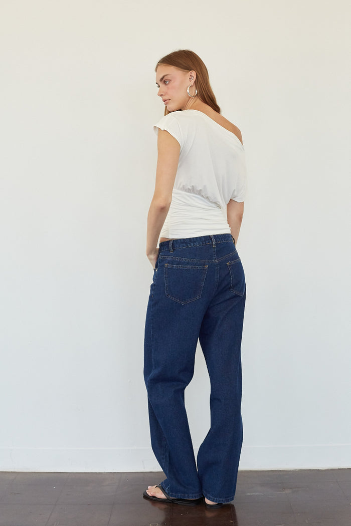 A woman stands in profile wearing dark blue wide-leg jeans, a white off-the-shoulder top, and flat sandals against a plain white wall.