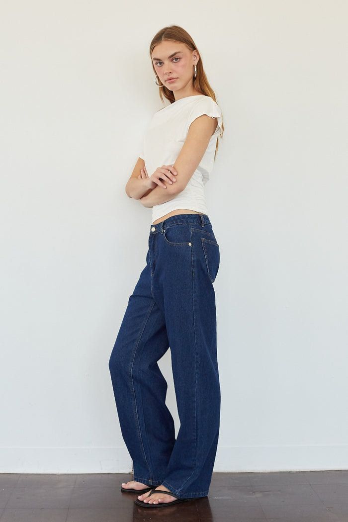A woman modeling a pair of dark blue wide-leg jeans with a white short-sleeve crop top, standing against a plain white wall.