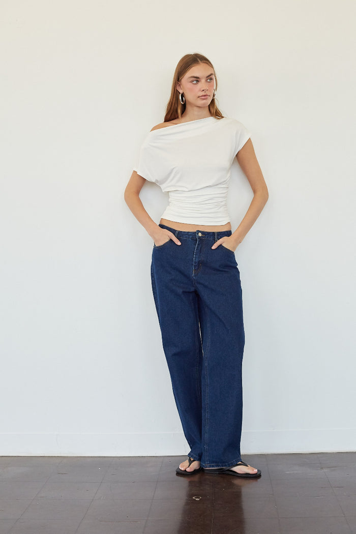 A woman stands against a plain white wall wearing a white off-the-shoulder wrap top and dark blue wide-leg jeans, with her hands in her pockets.
