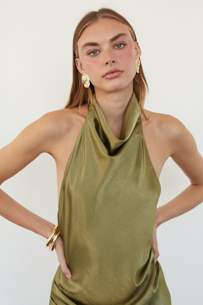 A woman modeling a sleeveless olive green halter-neck top with a draped cowl neckline, paired with gold earrings and a gold bangle, against a plain light background.