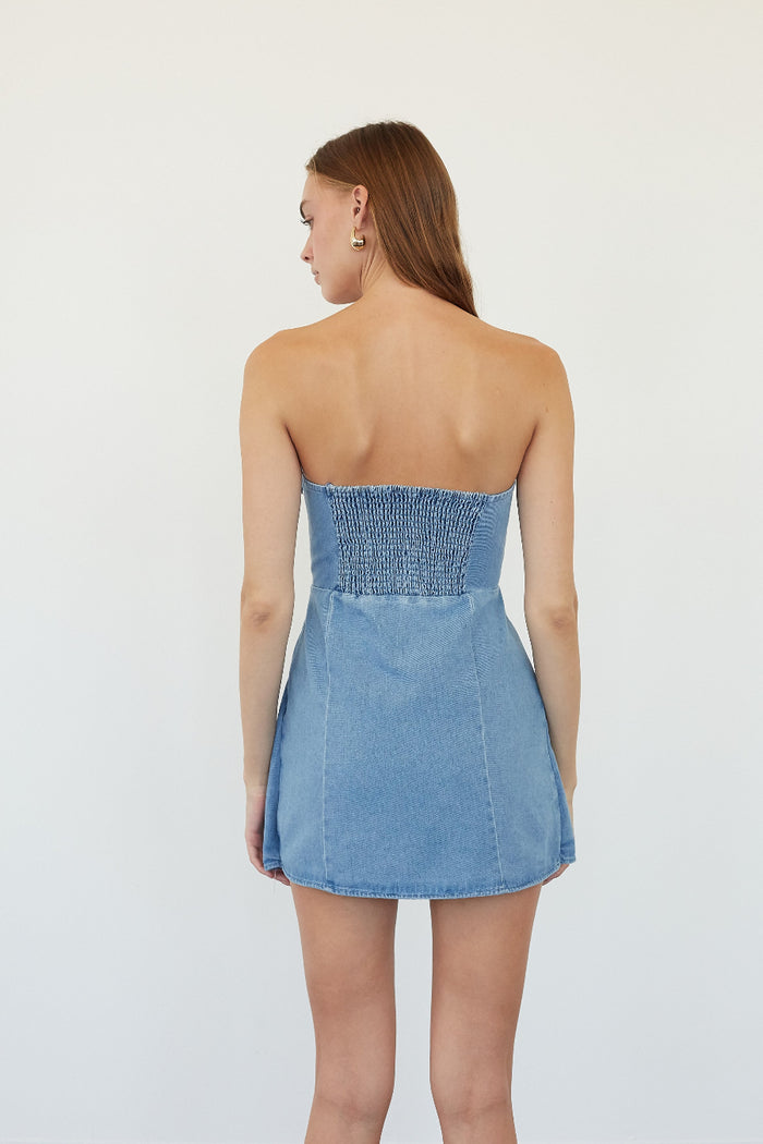 A woman shown from the back wearing a strapless light blue denim mini dress with a smocked bodice, set against a plain white studio background.