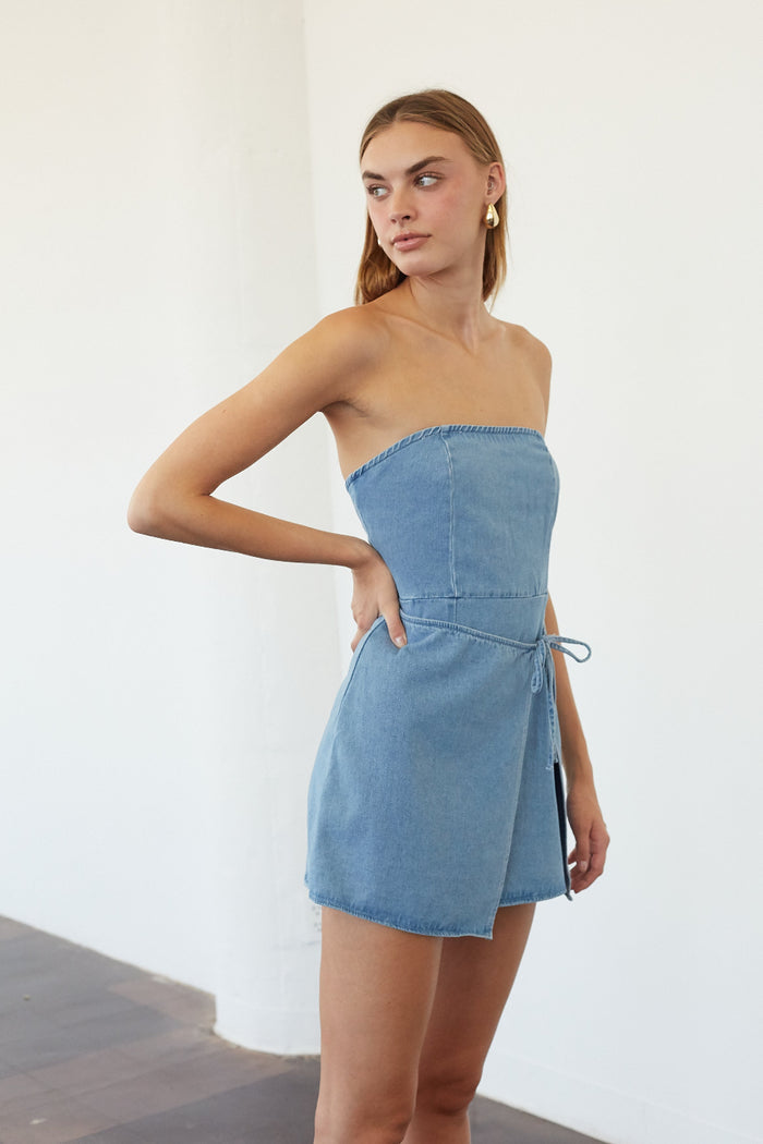 A woman modeling a strapless light blue denim mini dress with a tie detail at the waist, standing against a plain white wall.