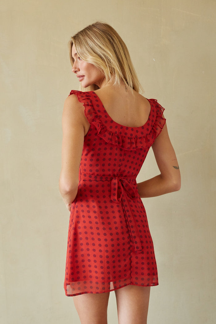 A woman wearing a red sleeveless dress with a black polka dot pattern, photographed from the back against a plain beige wall.