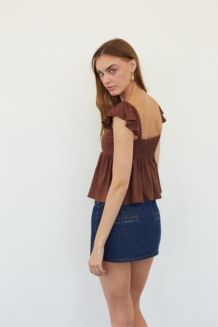A woman modeling a brown ruffled off-the-shoulder peplum top paired with a blue denim mini skirt, photographed against a plain white studio background.