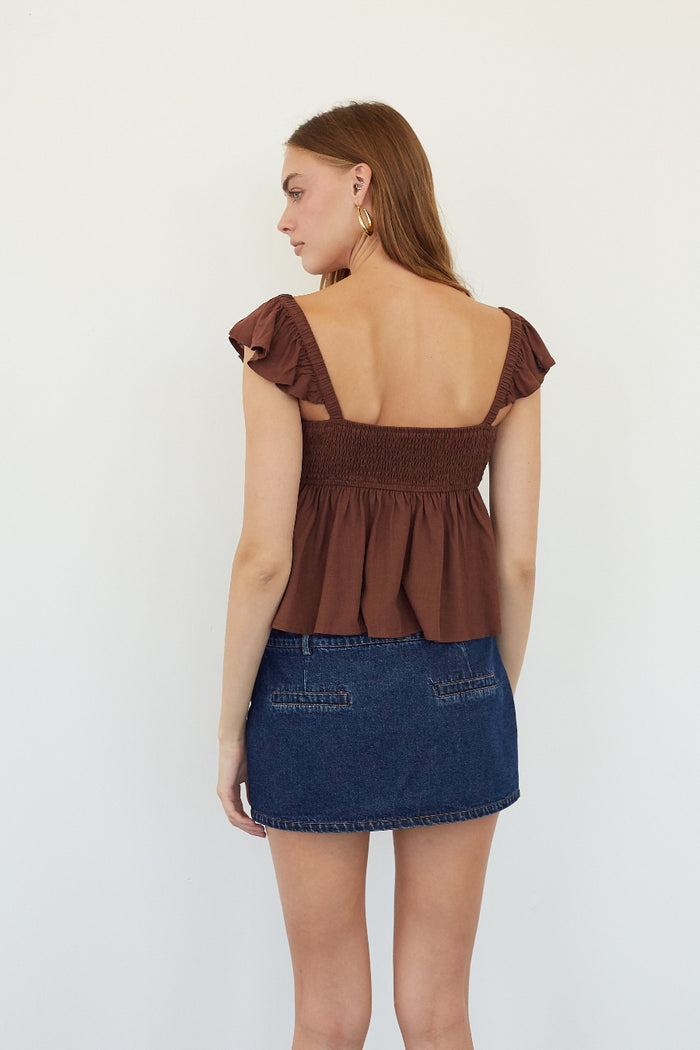 A woman shown from the back wearing a brown ruffled off-the-shoulder peplum top and a blue denim mini skirt, standing against a plain white background.