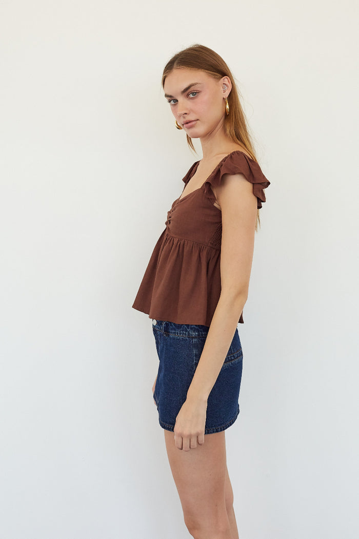 A woman modeling a brown ruffled sleeveless peplum top paired with denim shorts, standing against a plain white studio background.