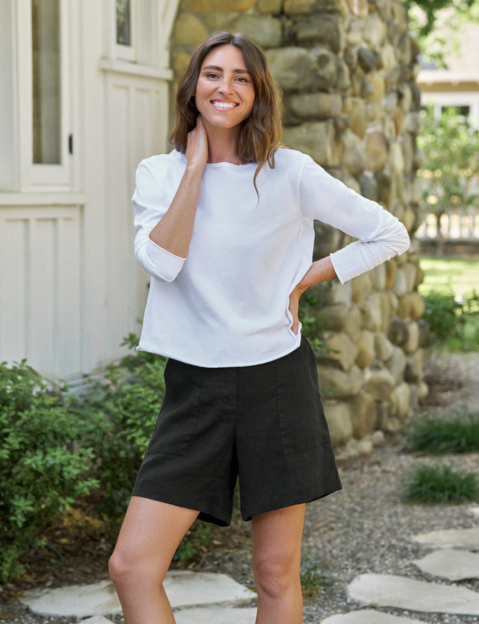 Woman wearing a white long-sleeve top and black shorts standing outdoors with a stone wall and greenery in the background.