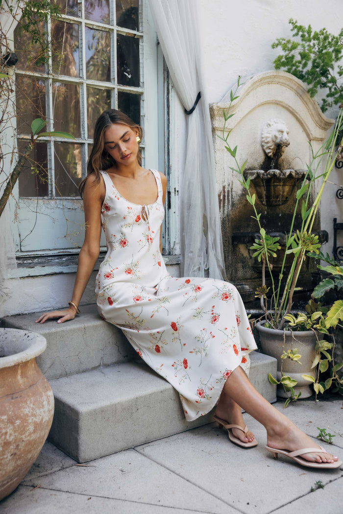 Woman in a floral dress sitting on steps outdoors with plants and a fountain in the background