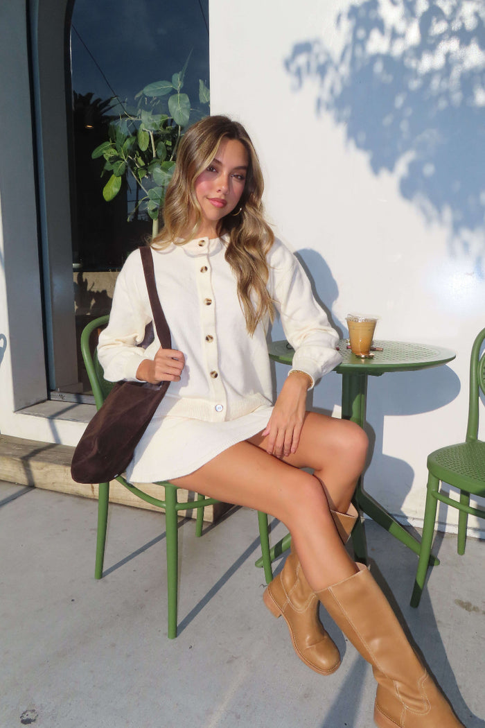 Woman sitting outdoors at a small table with a drink, wearing a white outfit and brown boots.