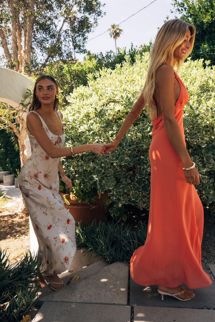 Two women in long dresses shaking hands outdoors with greenery in the background
