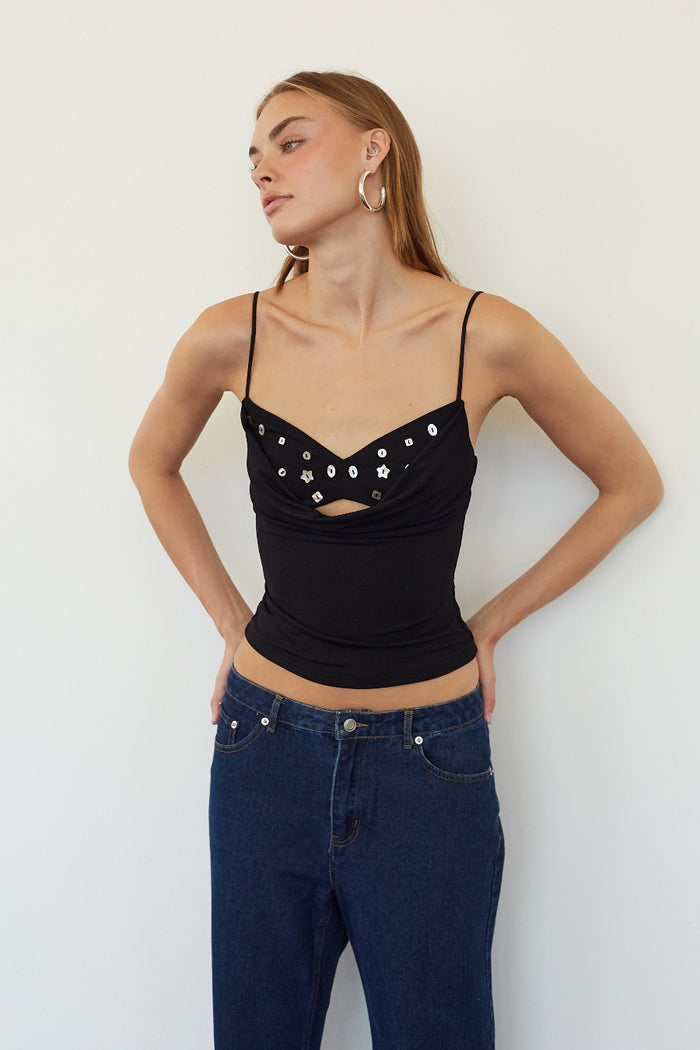 A woman modeling a black spaghetti-strap camisole top with silver rhinestone embellishments on the bust and a small keyhole cutout at the center, paired with dark blue jeans against a plain white wall.