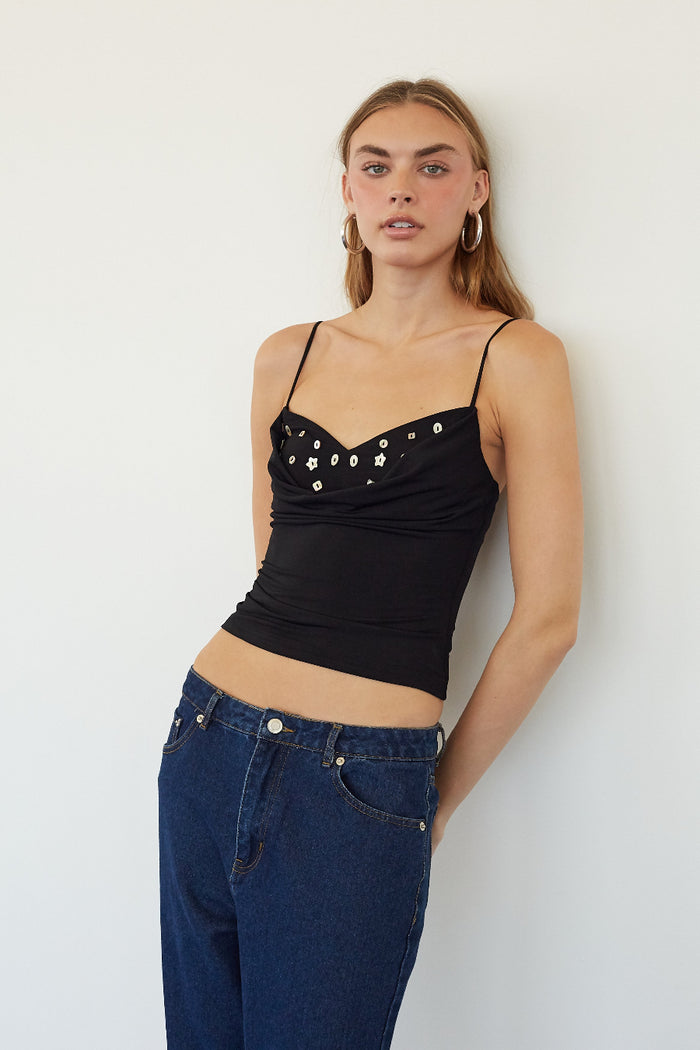 A woman modeling a black spaghetti-strap crop top with silver rhinestone embellishments on the bust, paired with dark blue jeans, standing against a plain white wall.