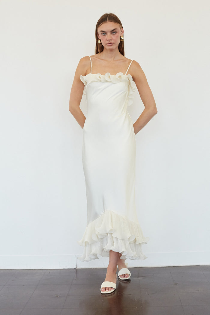 A woman modeling a white ruffled spaghetti-strap maxi dress with a fitted silhouette and a layered ruffle hem, standing against a white wall.
