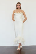 A woman modeling a white ruffled spaghetti-strap maxi dress with a fitted silhouette and a layered ruffle hem, standing against a white wall.