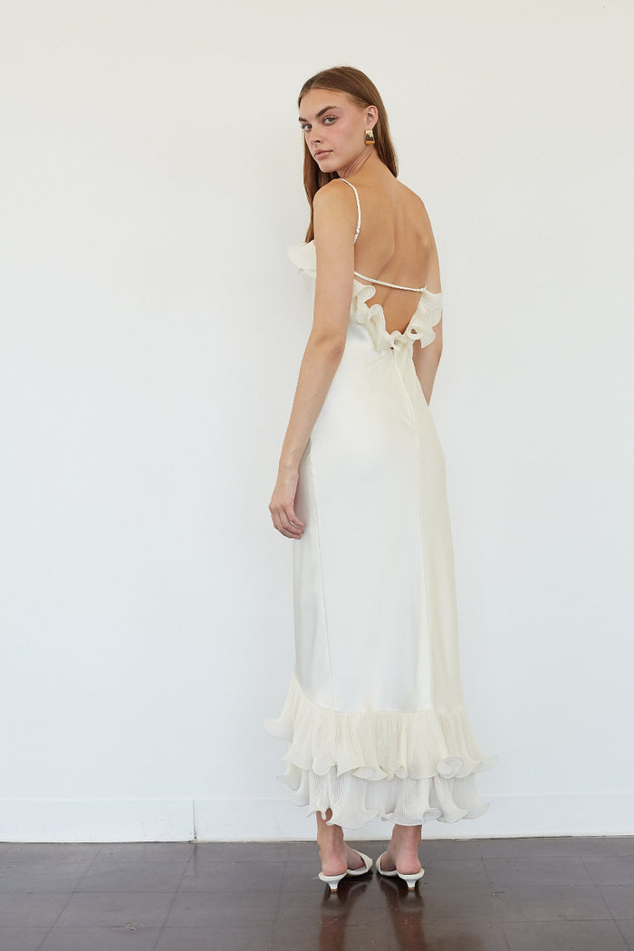 A woman modeling a long cream maxi dress from the back, featuring thin straps, a ruffled open back, and a tiered ruffle hem, set against a plain white wall.