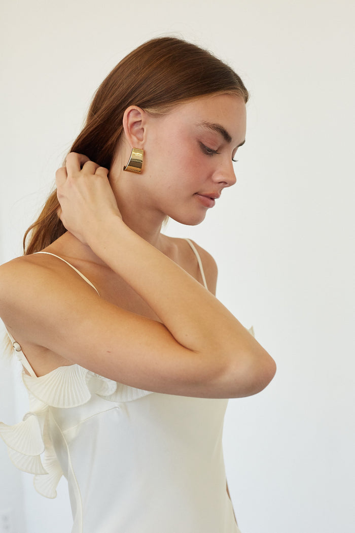 A woman wearing a white camisole and adjusting her hair, showing a chunky gold geometric dangle earring. The background is a clean, off-white studio wall.