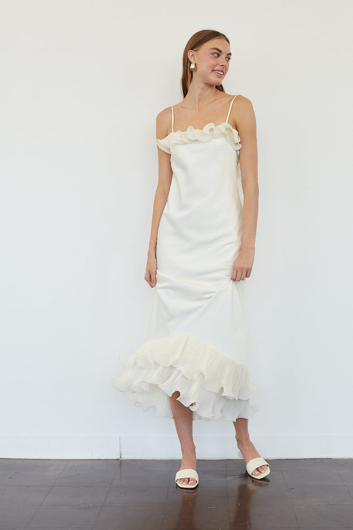 A woman modeling a white spaghetti-strap midi dress with ruffle details at the neckline and hem, paired with white slide sandals, standing against a plain white wall.