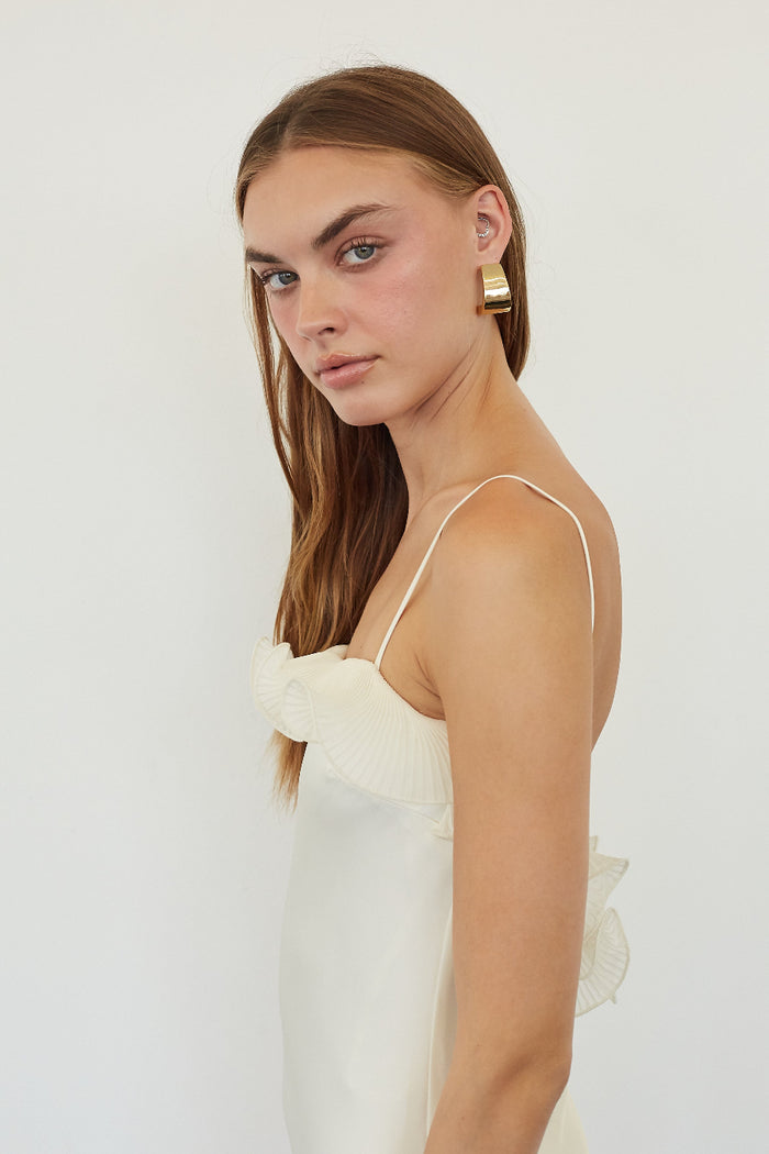 A woman with long light brown hair is shown in profile wearing a white spaghetti-strap top and a large gold geometric earring, set against a plain white studio background.