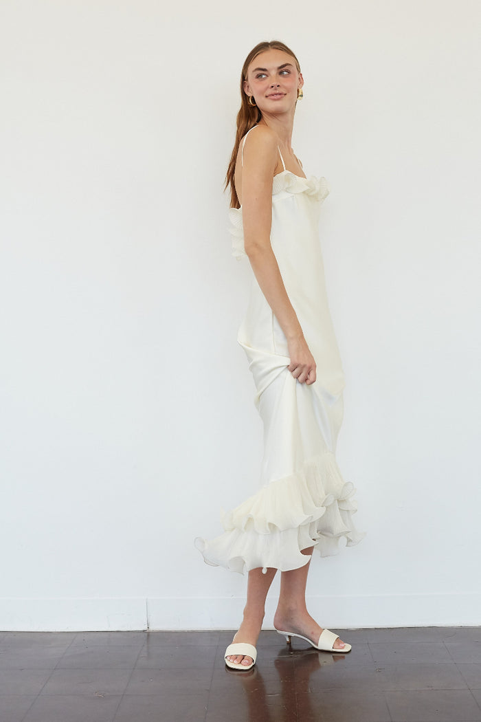 A woman modeling a white ruffled spaghetti-strap midi dress with a ruffled hem, paired with white slide sandals, standing against a white wall.